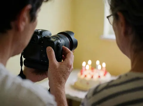 La main d'un photographe professionnel ajuste la mise au point de son appareil photo reflex numérique devant un gâteau d'anniversaire allumé dans une pièce aux murs jaunes.