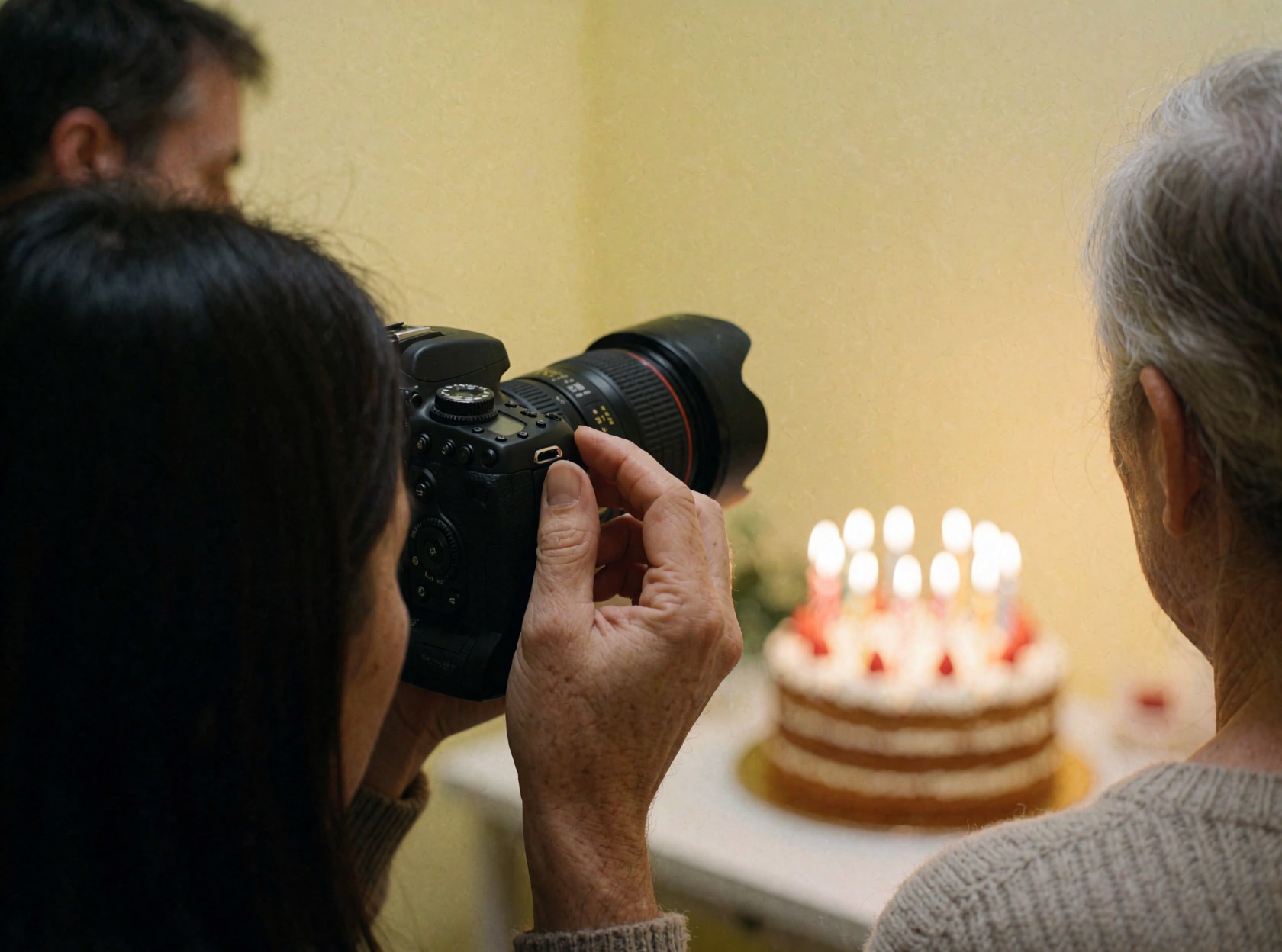 Une main ajuste l'anneau de mise au point d'un appareil photo reflex professionnel sur un gâteau aux bougies allumées dans un cadre chaleureux à murs jaunes.