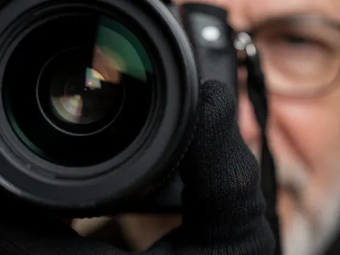 Gros plan d'une main portant un gant noir actionnant le bouton de prise de vue d'un objectif professionnel, avec un arrière-plan flou et des reflets lumineux dramatiques.
