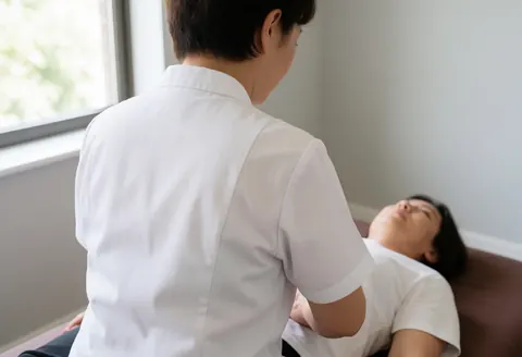Un praticien de shiatsu exerce une pression manuelle sur le ventre d'une cliente allongée sur un tapis dans une pièce baignée de lumière naturelle.