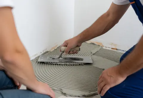 Un professionnel applique un adhésif gris sur un mur blanc à l'aide d'une truelle, dans le cadre de la pose d'un revêtement de sol en liège.