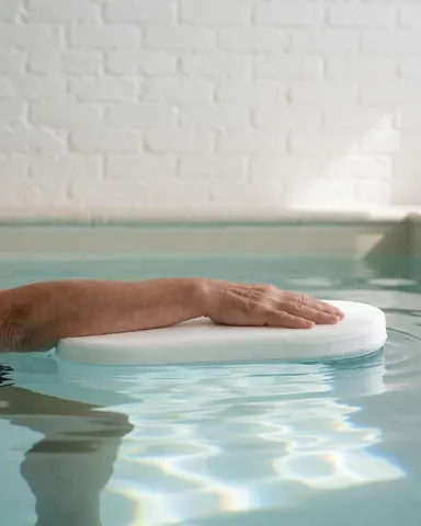 Une main adulte repose sur un panneau de natation flottant dans une eau claire avec des reflets solaires, devant un mur de briques blanches.