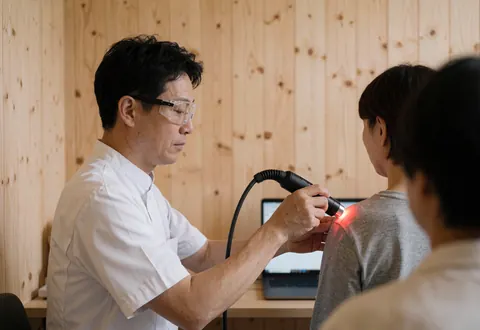 Un thérapeute portant des lunettes de sécurité utilise la lumière rouge pour traiter l'épaule d'un client dans un cabinet professionnel aux murs en bois clair.
