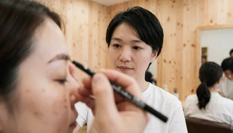 Une technicienne applique du maquillage dans un salon aux murs en bois, avec une cliente dont les sourcils sont visibles en premier plan.