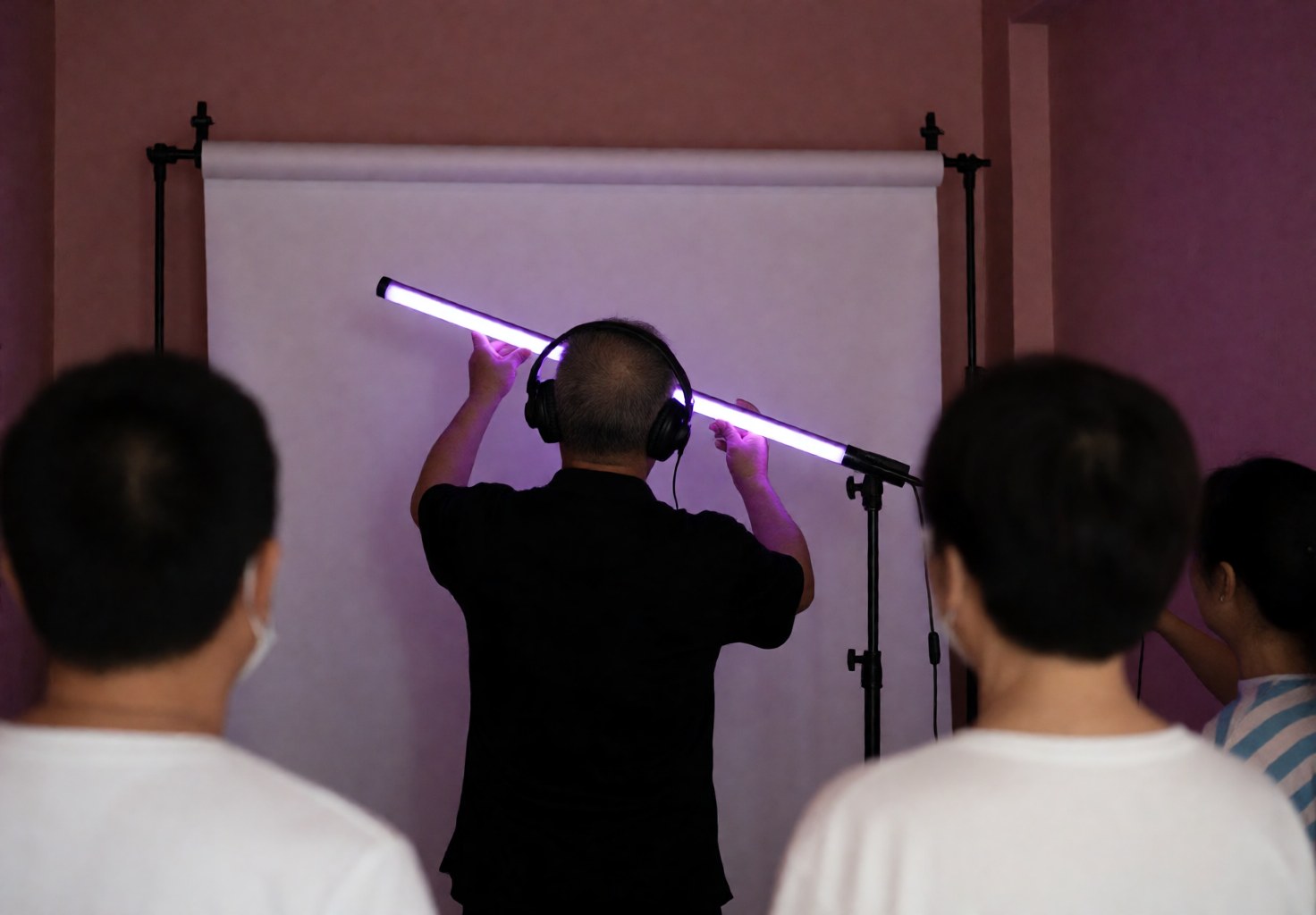 Photographe professionnel règle un éclairage LED violet devant un fond blanc dans un studio aux murs roses, des clients observent le service.