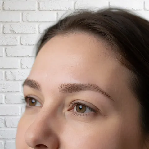 Gros plan sur un front lisse et des sourcils bien dessinés, éclairage doux sur un mur de briques blanches pour illustrer les soins capillaires.