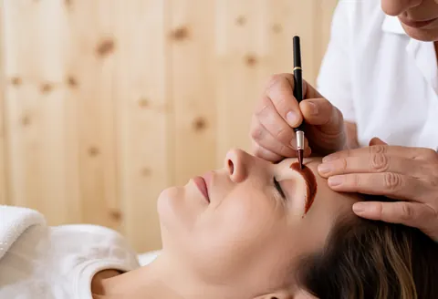 Une main utilise un pinceau fin pour appliquer du henné sur une sourcil dans un salon de beauté avec un mur en bois clair en arrière-plan.