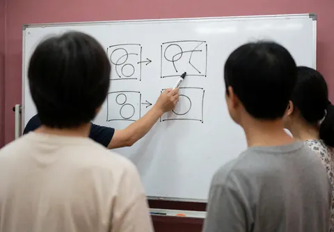 Un enseignant trace des schémas stratégiques sur un tableau blanc devant des élèves dans une salle de classe aux murs roses.