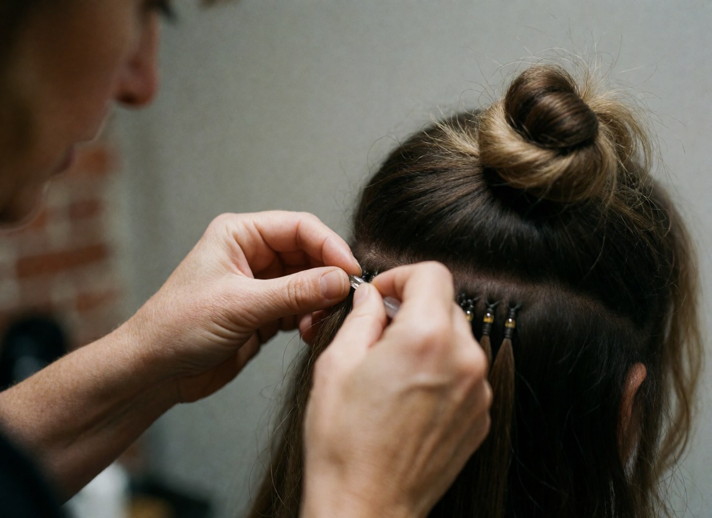 Des mains utilisent un outil pour fixer des extensions de cheveux ornées de perles devant un mur de briques rouges, illustrant un service de coiffure professionnel en cours.