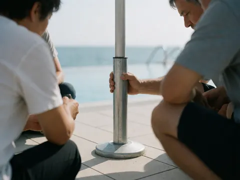 Deux personnes installent un grand parasol métallique sur la terrasse d'une piscine, avec une base lourde et un sol propre sous une lumière vive.