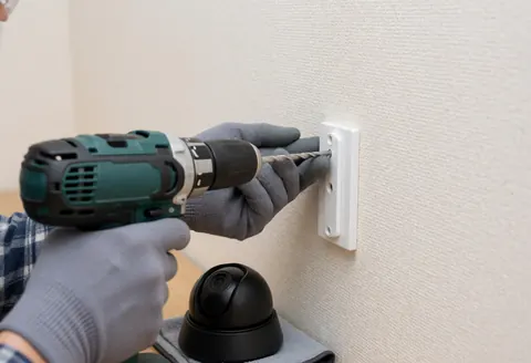 Un technicien portant des gants fixe une caméra de sécurité sur un mur beige, illustrant l'installation en cours pour un service professionnel.