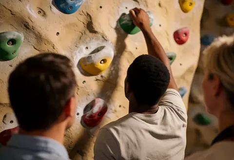 Un grimpeur atteint une prise verte sur un mur artificiel pendant que d'autres observent depuis le côté.