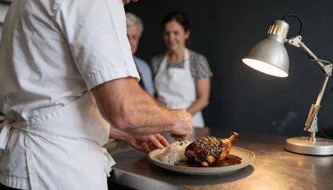 Un chef place des morceaux de poulet nappés de sauce mole sur un lit de riz blanc dans une assiette en céramique, le tout éclairé par une lampe à lumière chaude dans une cuisine aux murs sombres.
