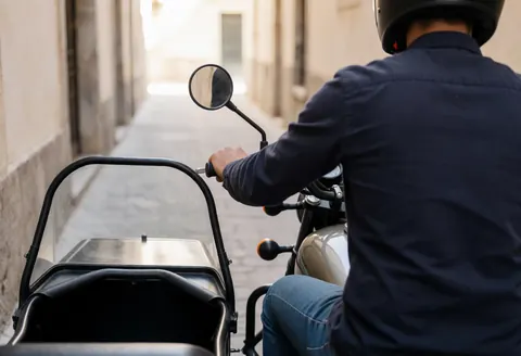 Conducteur de moto-taxi avec sidecar naviguant dans une rue étroite, main sur le guidon, murs de bâtiment, service en action.