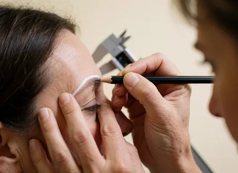Un professionnel trace une ligne blanche avec un crayon sur le sourcil d'une cliente, un calibreur métallique visible en arrière-plan dans un salon de microblading.