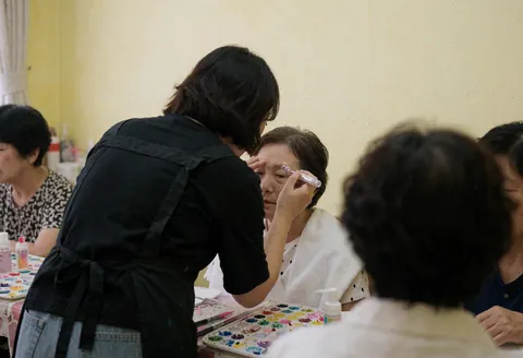 Artiste professionnel appliquant du glitter sur l'œil d'une cliente assise, avec palette de couleurs et murs jaunes clairs dans un salon de maquillage.