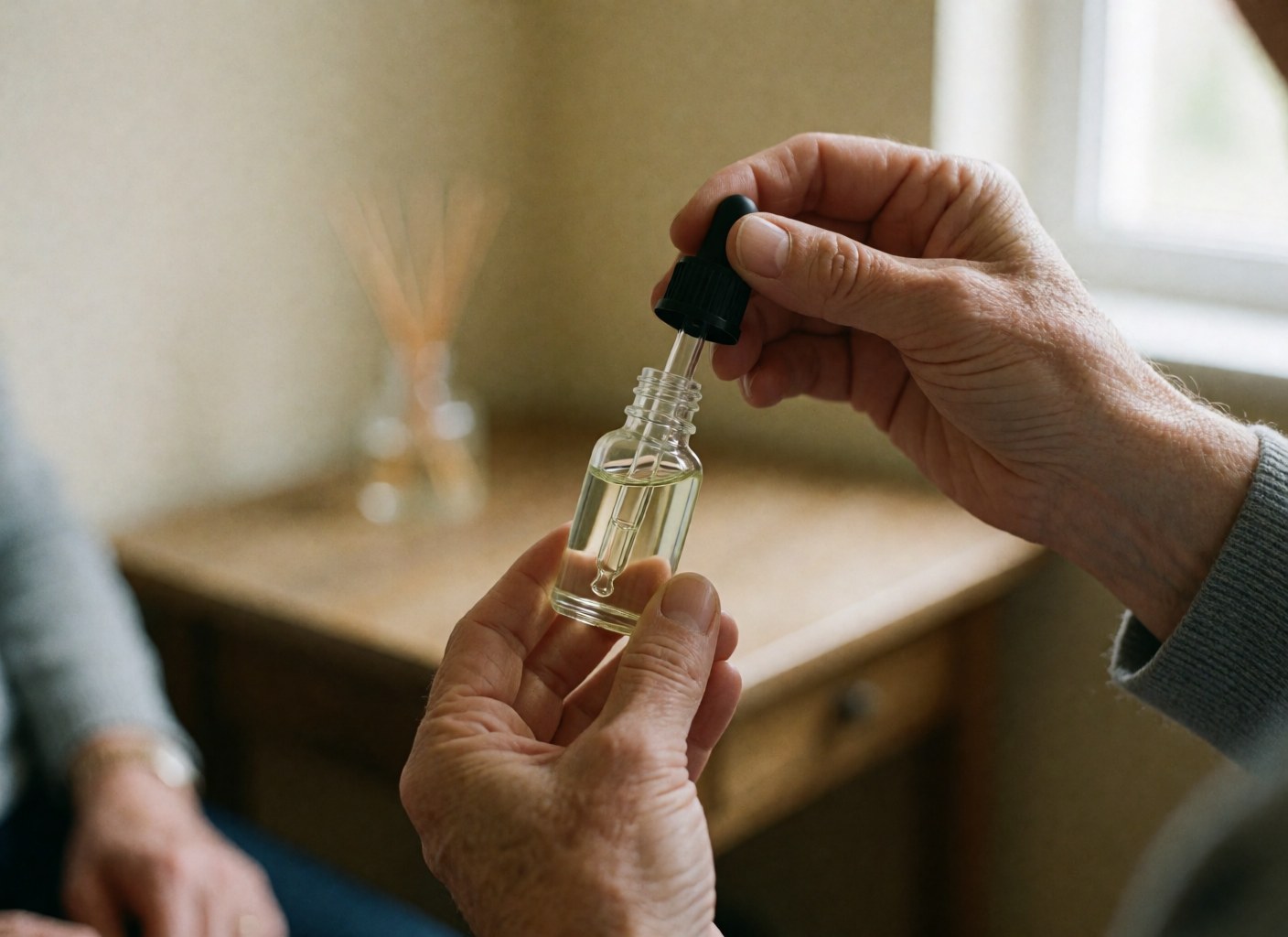 Des mains adultes tiennent un flacon en verre avec un compte-gouttes noir, devant un diffuseur en bois, baigné d'une lumière douce de fenêtre.