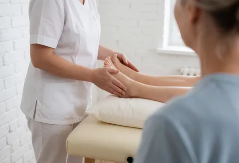 Un thérapeute réalise un massage de drainage lymphatique sur le pied d'une cliente dans une salle avec mur en briques blanches et lumière naturelle.