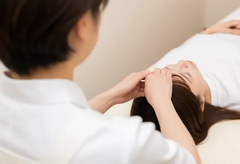 Un thérapeute applique un massage sur la tête d'une cliente allongée sur une table de massage, dans un cadre professionnel avec des vêtements blancs et un fond neutre.