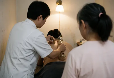 Un thérapeute applique des ventouses de cupping sur le dos nu d'un client allongé, tandis qu'une autre personne observe la scène dans un cadre professionnel et chaleureux.