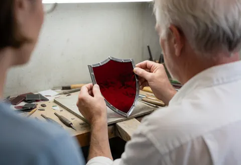 Un artisan assemble un blason avec du verre rouge dans un atelier de cristallerie décorative, outils visibles.
