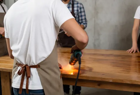 Un technicien portant un tablier utilise un appareil de chauffage sur une table en bois dans un atelier avec un mur en béton et de la fumée visible.
