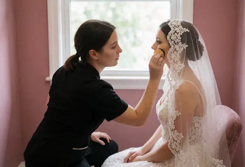 Une maquilleuse professionnelle applique de la base sur le visage d'une mariée portant un voile brodé dans une pièce aux murs roses et à la lumière naturelle.