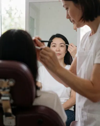 Une cliente observe ses sourcils traités dans un miroir pendant qu'une technicienne applique le produit en salon de beauté.