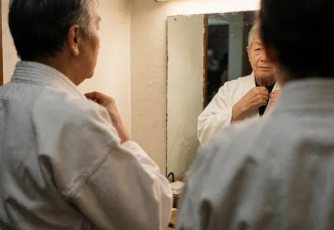 Une personne ajuste le col de son uniforme de karaté blanc devant un miroir dans un dojo aux murs clairs, mettant en valeur les détails du vêtement.