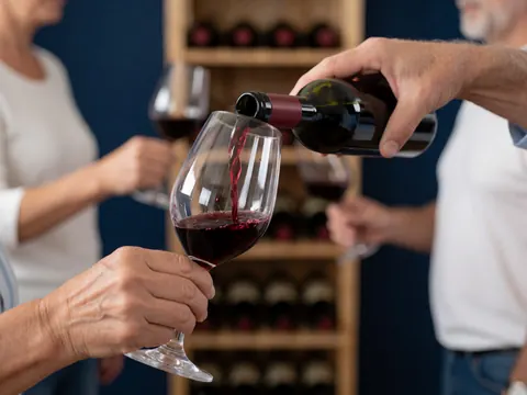 Une main verse du vin rouge dans un verre devant une étagère en bois et un mur bleu foncé, idéal pour une boutique en ligne de vins.