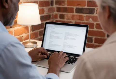 Une personne utilise un ordinateur portable sur un bureau devant un mur de briques, éclairage chaleureux et clavier visible.