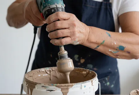 Une main utilise un malaxeur électrique pour mélanger de la peinture épaisse dans un seau, illustrant un service de peinture en cours.