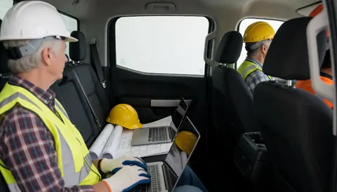 Deux ouvriers en casques et gilets de sécurité utilisant des ordinateurs portables et des plans à l'intérieur d'une camionnette.