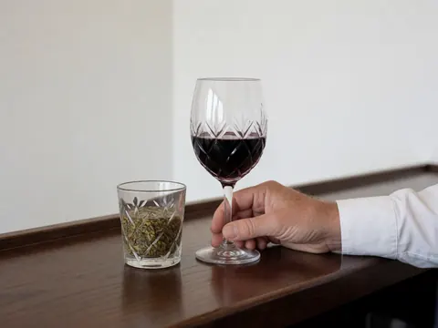 Une main tenant un verre de vin rouge à côté d'un tumbler avec des herbes sur un comptoir en bois poli devant un mur blanc.