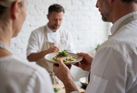 Un cuisinier en train de servir un plat de poisson avec des herbes dans un atelier de cuisine aux murs de briques blanches.