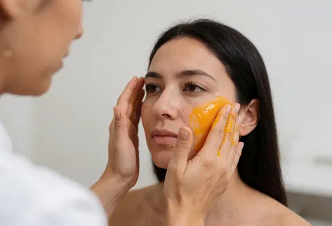Un praticien applique délicatement un masque hydratant à l'orange sur le visage d'une cliente dans un salon de beauté au fond neutre.