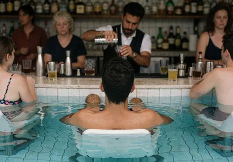 Un bartender sert une boisson à des clients près d'une piscine, avec une barre visible en arrière-plan.