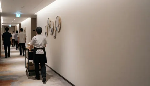 Un agent de nettoyage pousse un charrier dans le couloir d'un hôtel, avec de l'art mural et une lumière chaude.