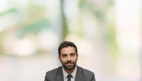 Un homme d'affaires portant un costume et une cravate regarde directement la caméra sur un fond flou de bureau moderne.