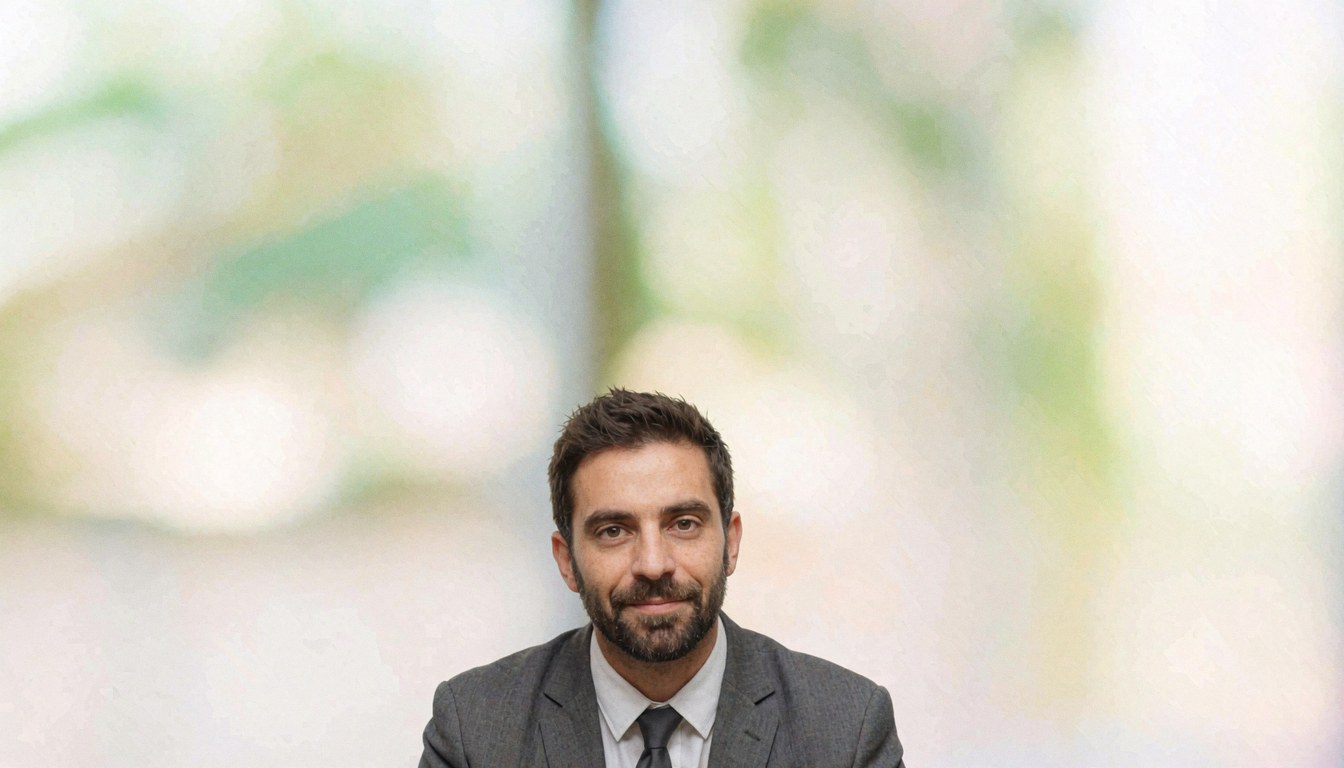 Un homme d'affaires portant un costume et une cravate regarde directement la caméra sur un fond flou de bureau moderne.