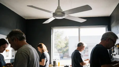 Technicien réparant un ventilateur blanc dans un atelier aux murs gris avec outils suspendus et éclairage naturel.