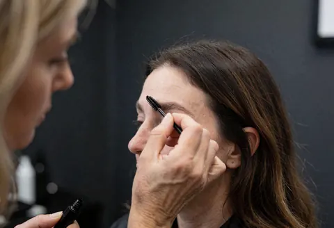Un spécialiste applique du gel sur les sourcils d'un client dans un salon de beauté avec un mur gris et des outils visibles.