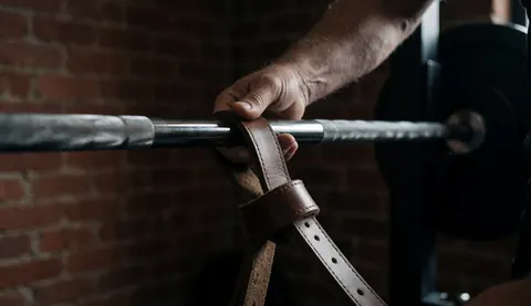 Gros plan d'une main liée par des sangles en cuir autour d'une barre de haltère dans un gymnastique aux murs de briques, illustrant l'entraînement de force.