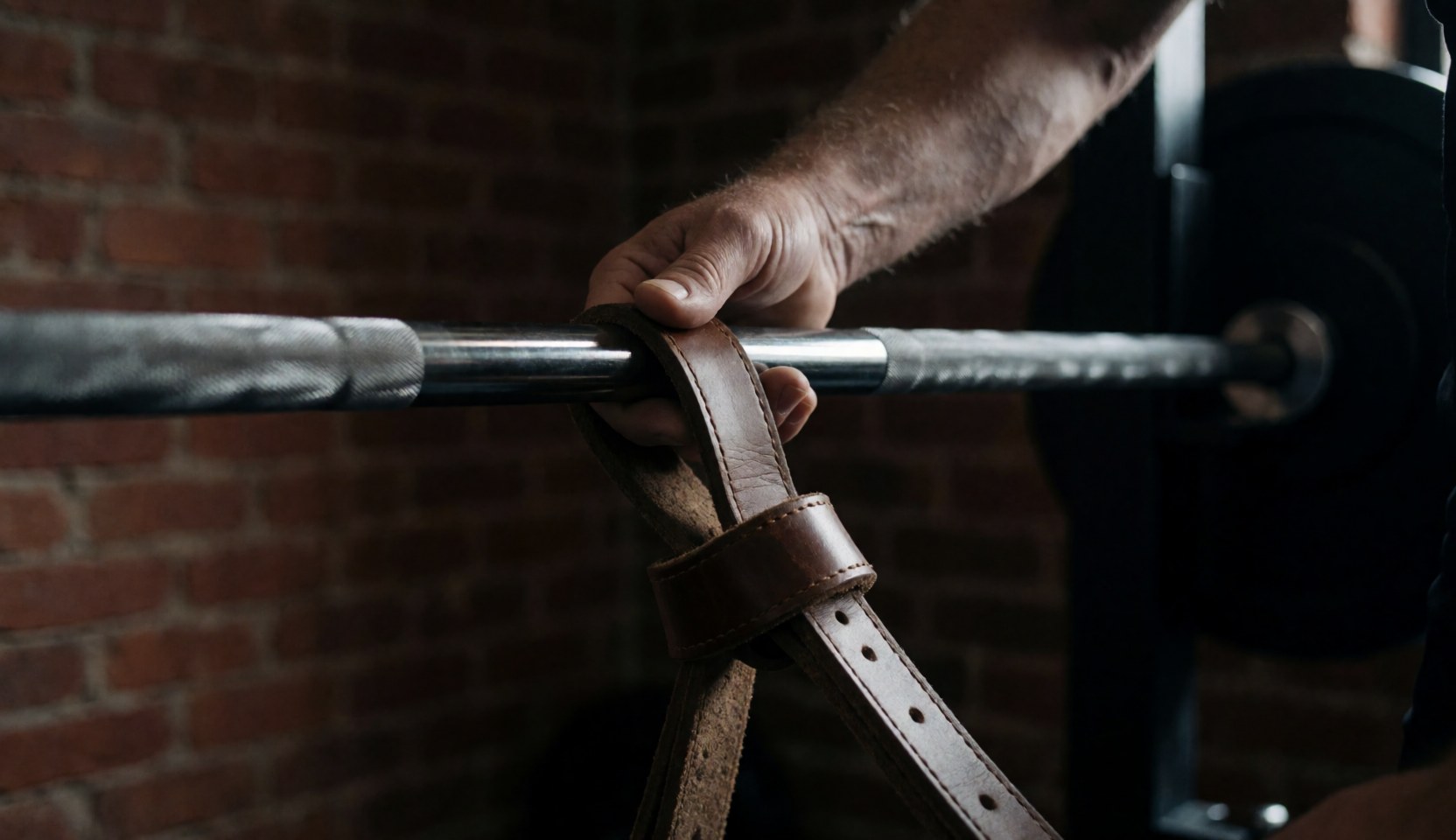 Gros plan d'une main liée par des sangles en cuir autour d'une barre de haltère dans un gymnastique aux murs de briques, illustrant l'entraînement de force.