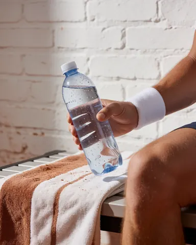 Une main tenant une bouteille d'eau avec des gouttes de condensation, posée sur une serviette devant un mur de briques blanches, évoquant le repos après le sport.