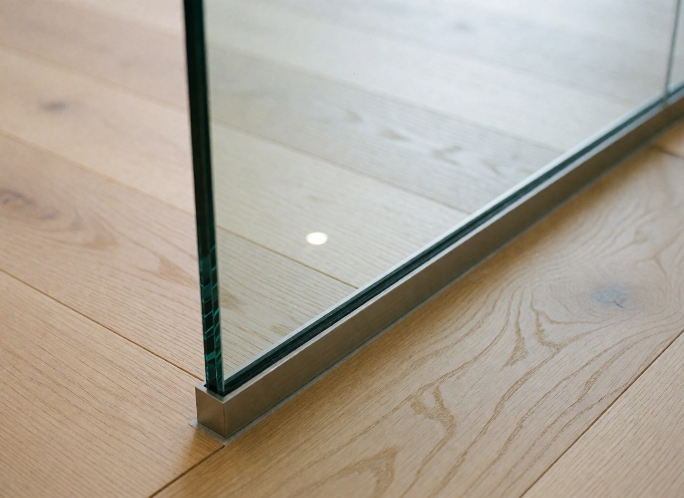 Détail d'un mur en verre sans cadre dans une clinique, avec un sol en bois clair et une plinthe en acier inoxydable, montrant un reflet de lumière.