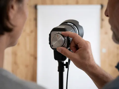 Une main ajuste un dial de lumière sur un équipement photographique professionnel devant un fond blanc et une paroi en bois clair.