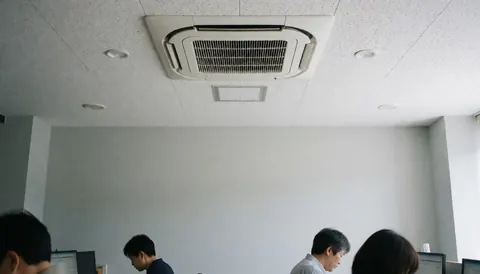 Vue d'un bureau moderne avec une unité de climatisation cassette au plafond, des employés travaillant sur des ordinateurs et des murs gris clair, éclairage naturel.