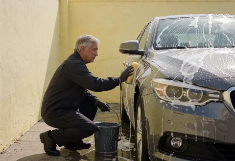 Un homme lave une voiture à l'aide d'une éponge et d'un seau dans une cour pavée, illustrant le service de nettoyage automobile en action.