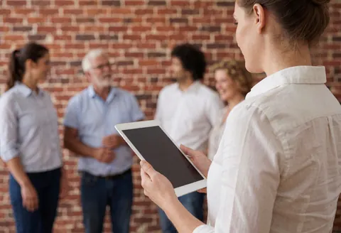 Une femme professionnelle tient une tablette tout en s'entretenant avec un groupe de personnes dans un espace intérieur doté d'un mur en briques apparentes.